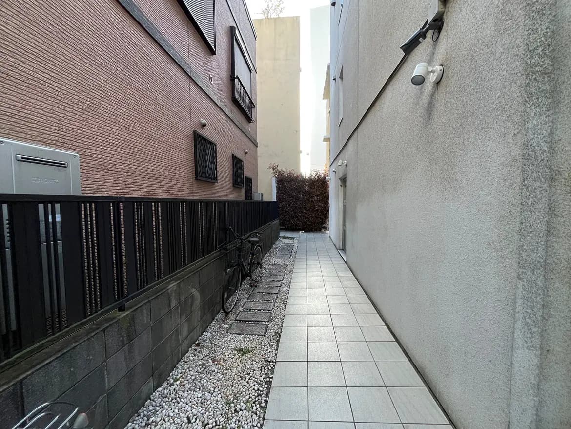 - 薄暗い通路に並ぶ灰色のタイル、左右には住宅の外壁とフェンス、奥には茂った植栽が見える風景