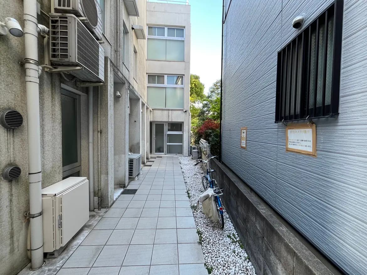 - 鉄筋コンクリートの建物の間を通る細い歩道、エアコンと窓が並ぶ、白い砂利が敷かれた小道の風景