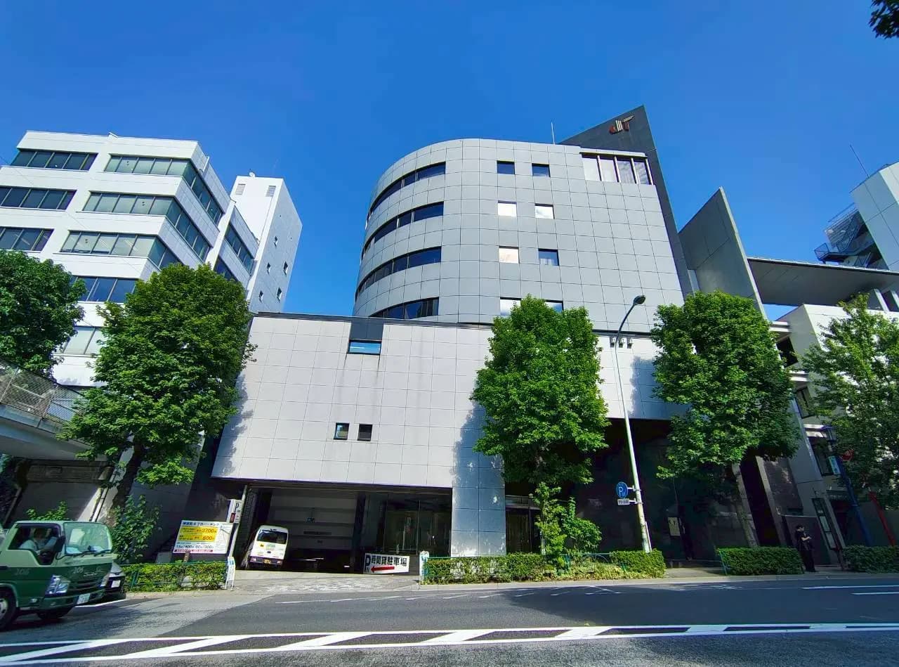 - 現代的なデザインのグレーの建物、周囲に緑の木々と青空の下での景観、駐車場と交通標識が見える通りの風景