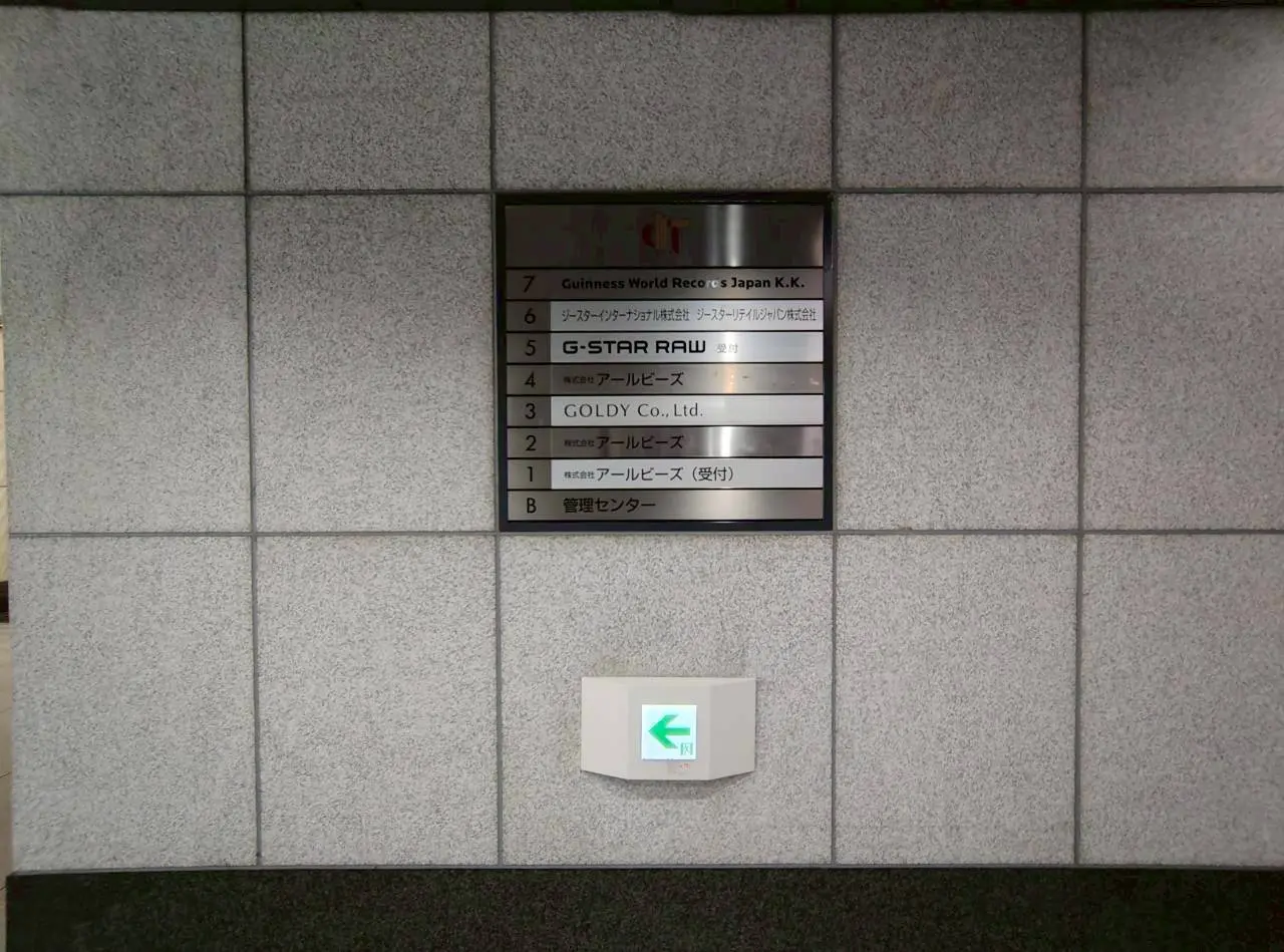- 企業名が表示された案内板、グレーのタイル壁に取り付けられたインフォメーションサイン、エレベーターや管理センターの表示も含む