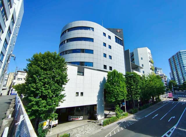 - 近代的な外観のビル、周囲に緑豊かな木々が並び、青空の下に広がる都市の風景