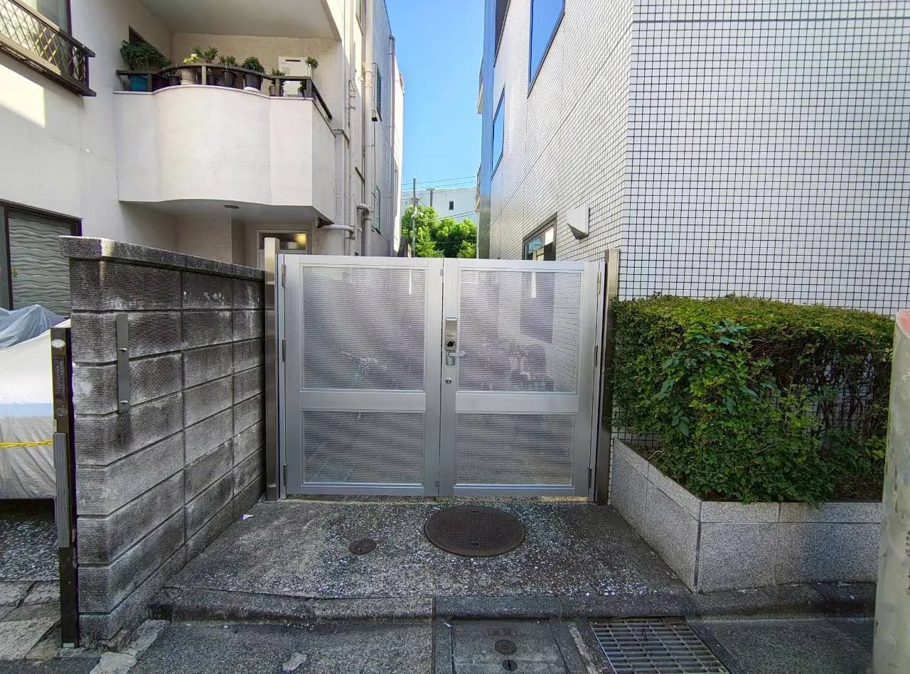 - 鉄製のメッシュゲートがある入り口、隣接する建物とグリーンの生垣が見える都市の景観