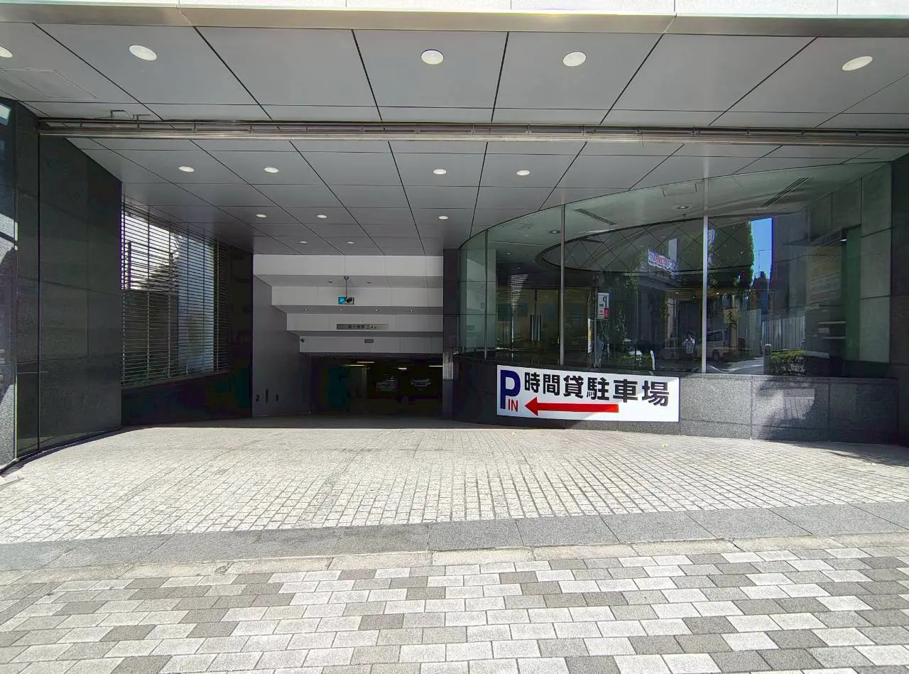 - 時間貸駐車場の入口、周囲のモダンな建物と整然とした石畳、左側に駐車場への案内看板がある風景