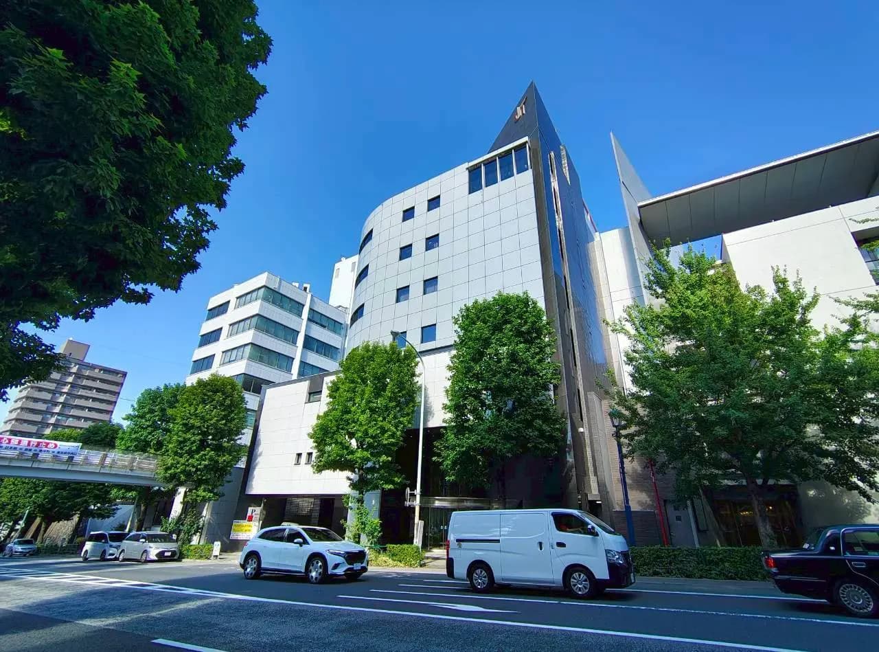 - 近代的なオフィスビルの外観、緑豊かな街並みと青空が広がる風景、車が走る通りの風景