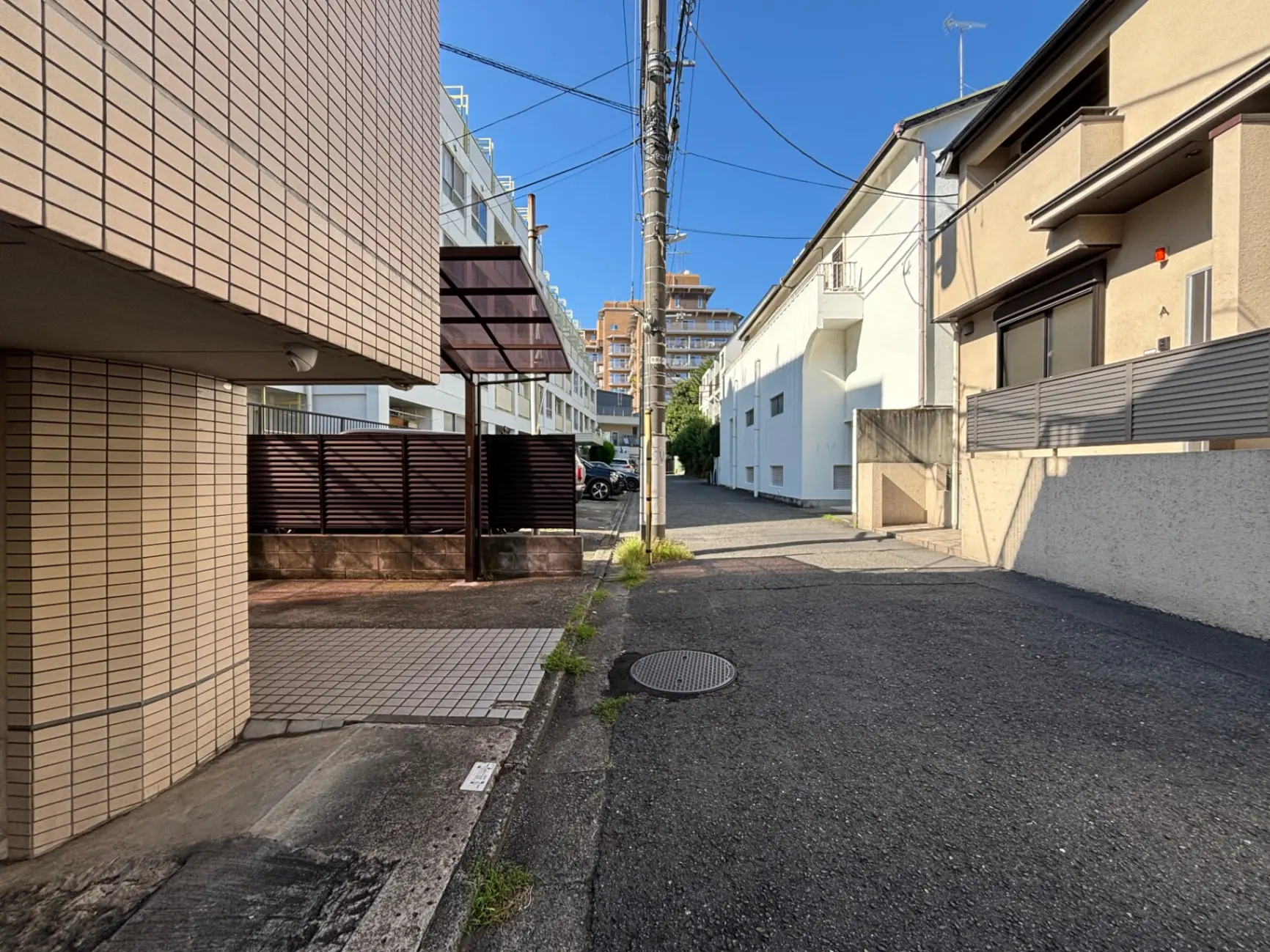 - 街角の道路と建物、青空の下に見える住宅と駐車場の風景、コンクリートとタイルの舗装が確認できる通りの様子