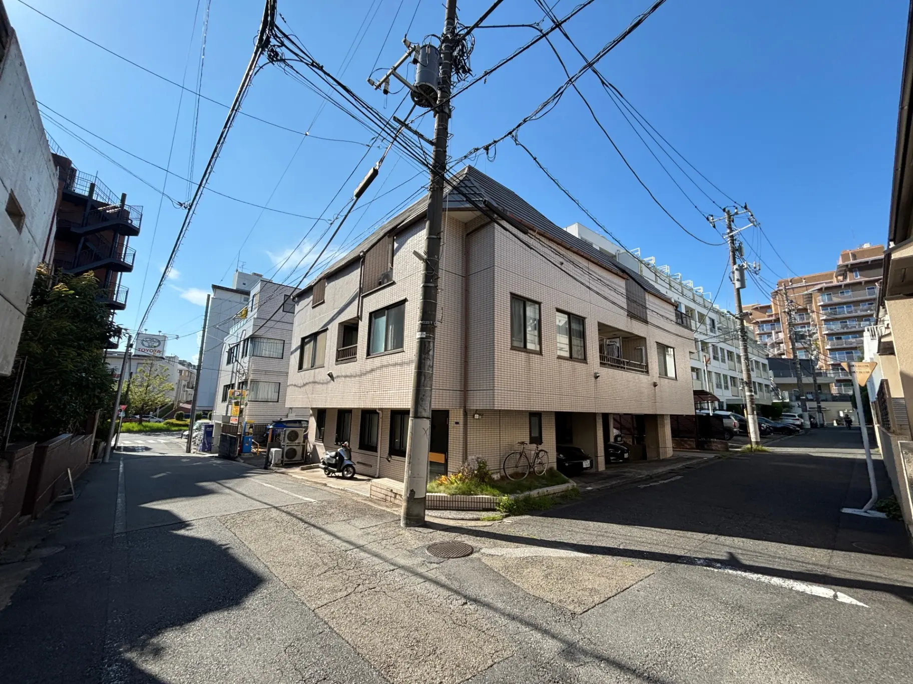 - 明るい青空の下で、住宅街の路地とコンクリートのアパート、電柱と電線が見える風景