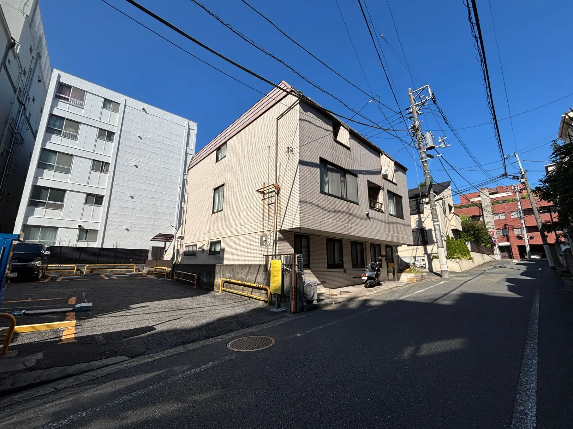 - 都市の住宅街にある三階建ての建物、青空の下で電線が走る風景、駐車場が見える通りの様子