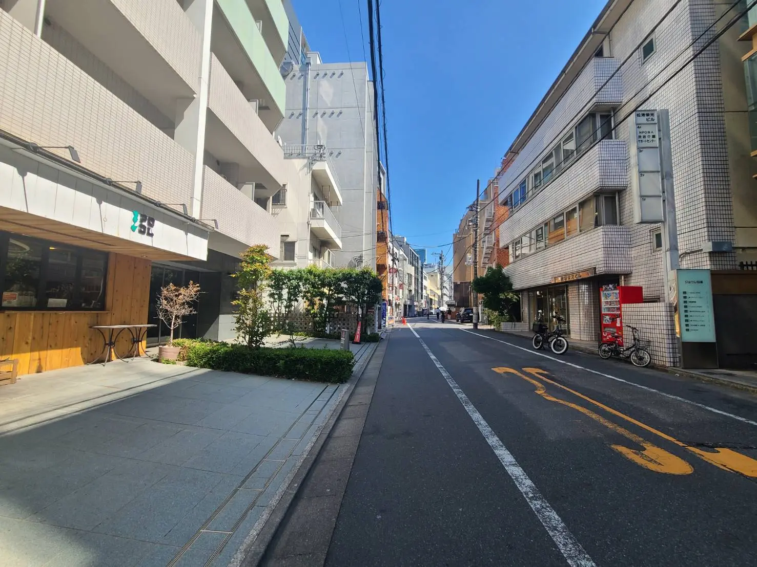 - 住宅街の通り、直線的な道路と両脇に並ぶ建物、青い空と明るい日差しが差し込む風景、手前にテーブルと植物のあるカフェのエントランス