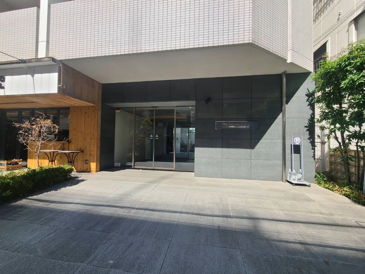 - モダンなデザインのエントランス、ガラス扉と石材の壁、木製のアクセントと緑の植栽で囲まれたエントランスホール