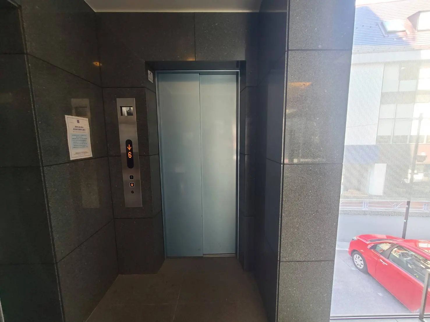 - モダンなエレベーターの内部、銀色のボタンパネルと透明な扉、隣には車が見える窓のあるエレベーターホール