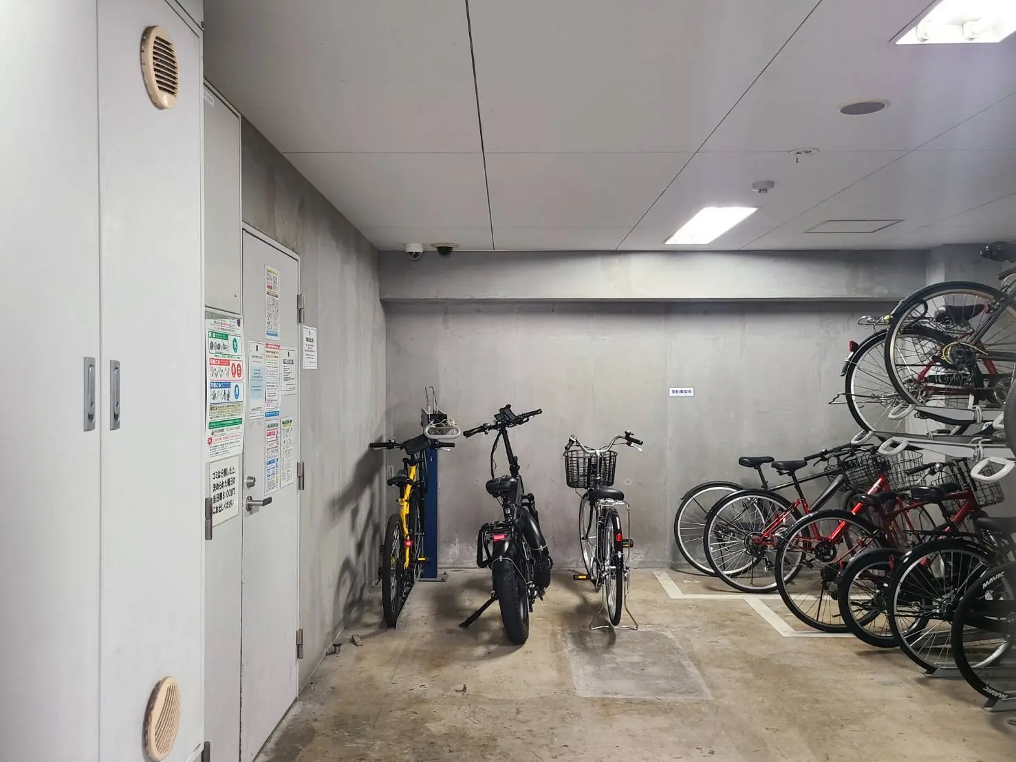 - コンクリート壁の自転車置き場、様々な色の自転車が整然と並ぶ風景、壁に貼られた注意事項の貼り紙がある室内