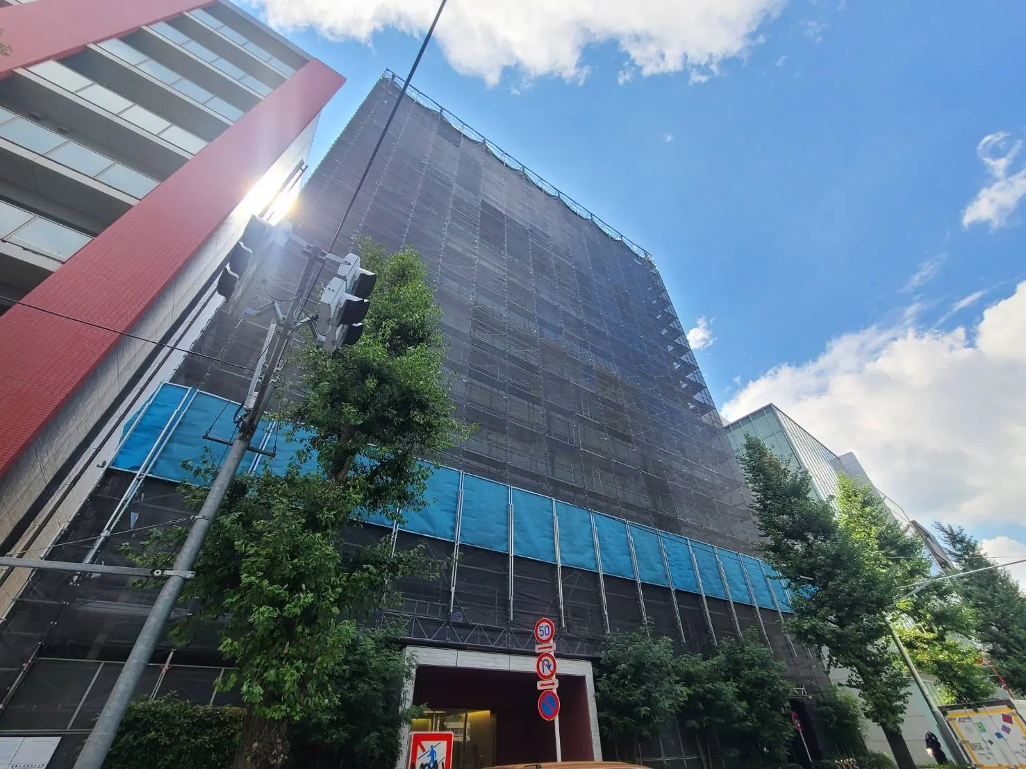 - 修繕中の高層ビル、青いシートで覆われている外観と周囲の緑、青空が広がる晴れた日の風景