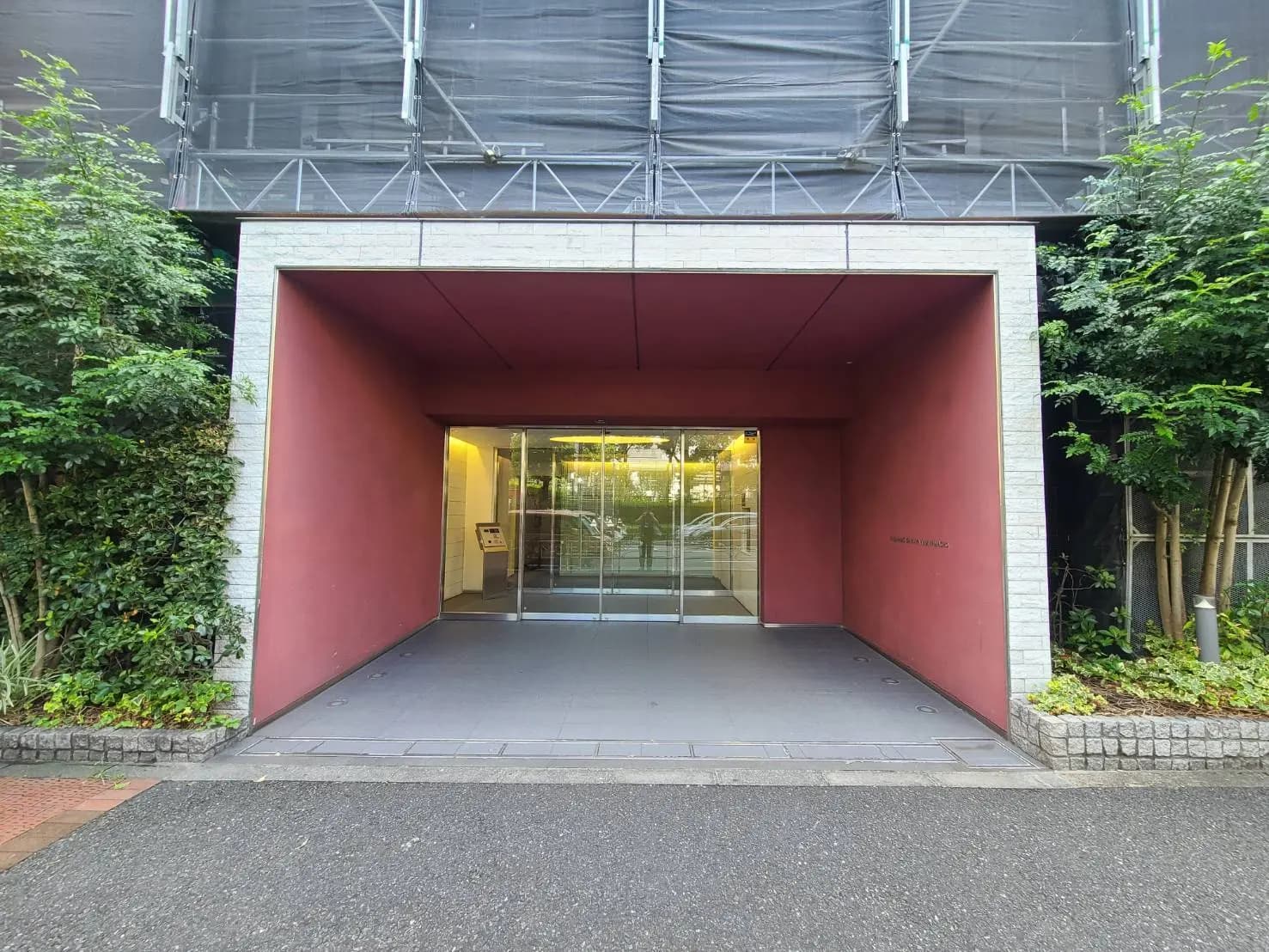 - 赤い壁のエントランス、ガラスの自動ドアと周囲の緑が調和するモダンな建物の入口