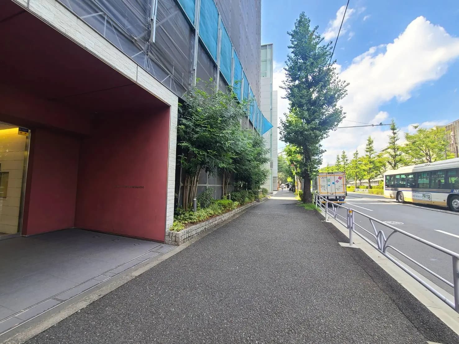 - 住宅街の歩道脇、緑豊かな植栽を背景にした赤い外壁の建物と明るい青空、通りに停車中のバスとトラックが見える風景