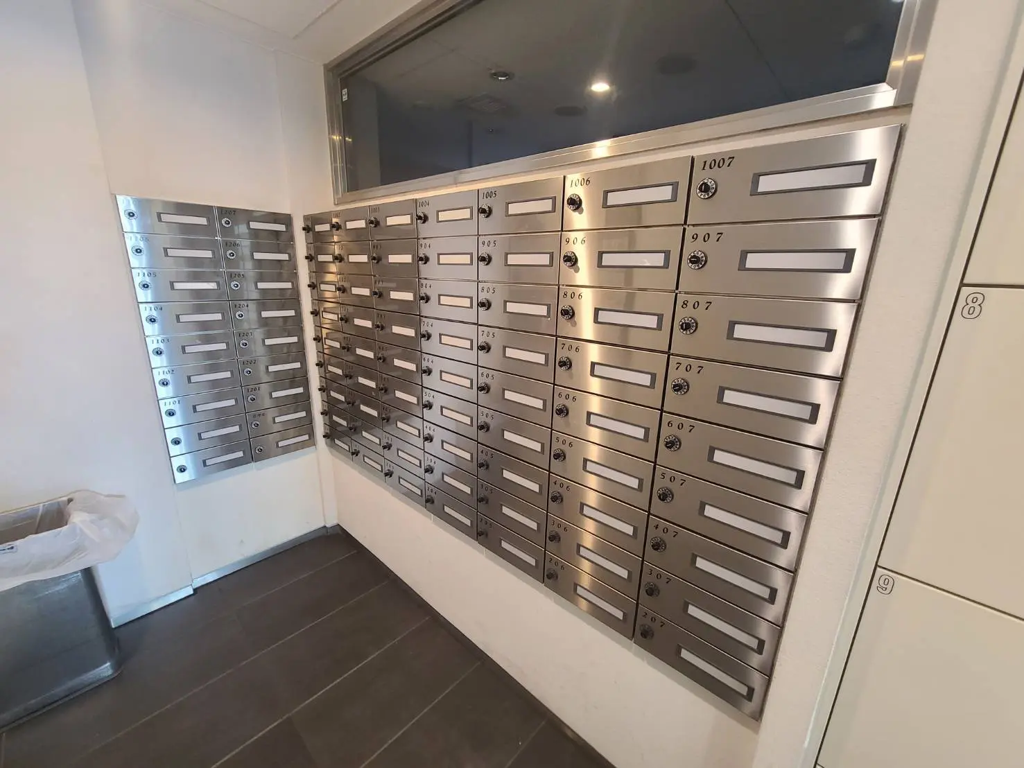 - ステンレス製の郵便受けが並ぶ現代的なロビー、壁に設置されたダイレクトメールボックスとゴミ箱も見えるインテリア