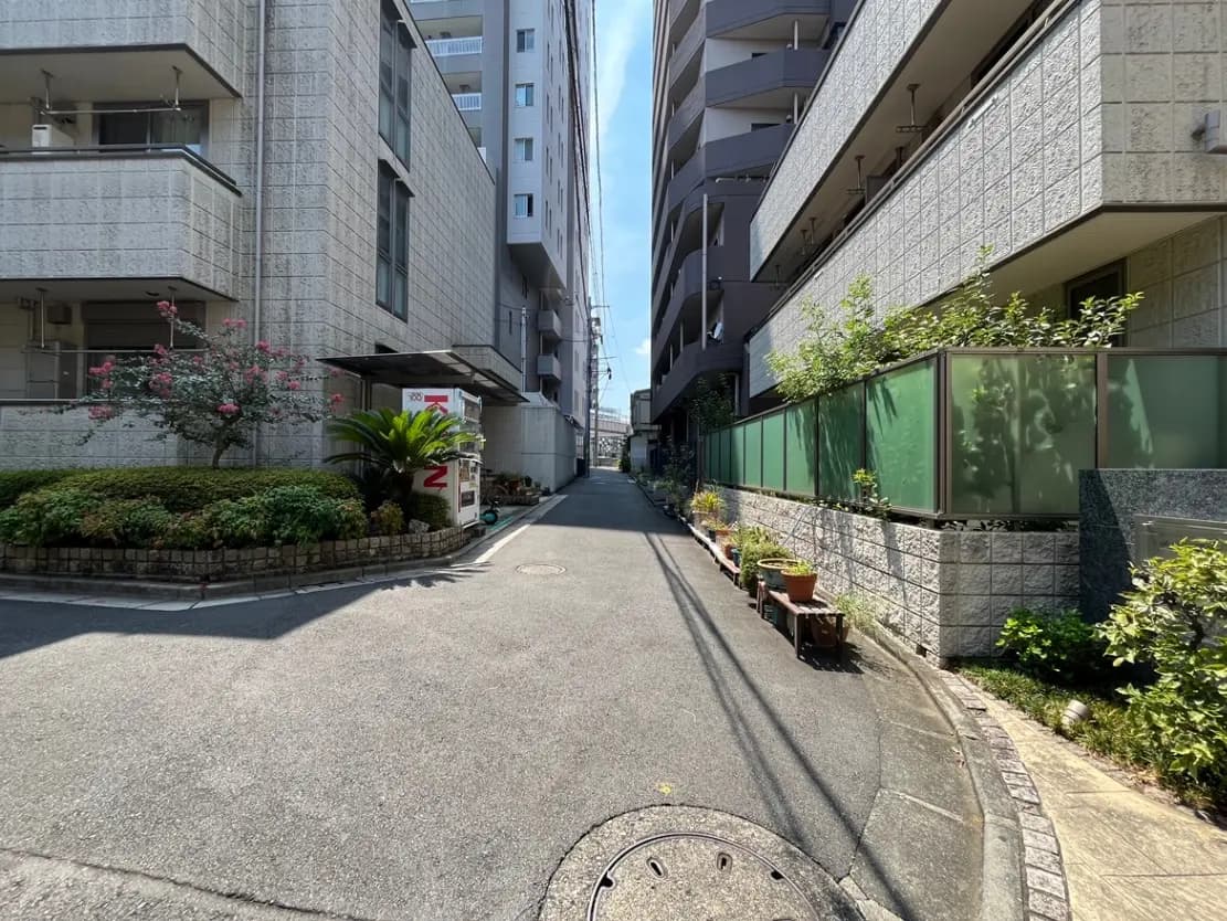 - 都市部の住宅街、マンションの隣に並ぶ緑のフェンスと植栽、明るい青空の下の静かな道路