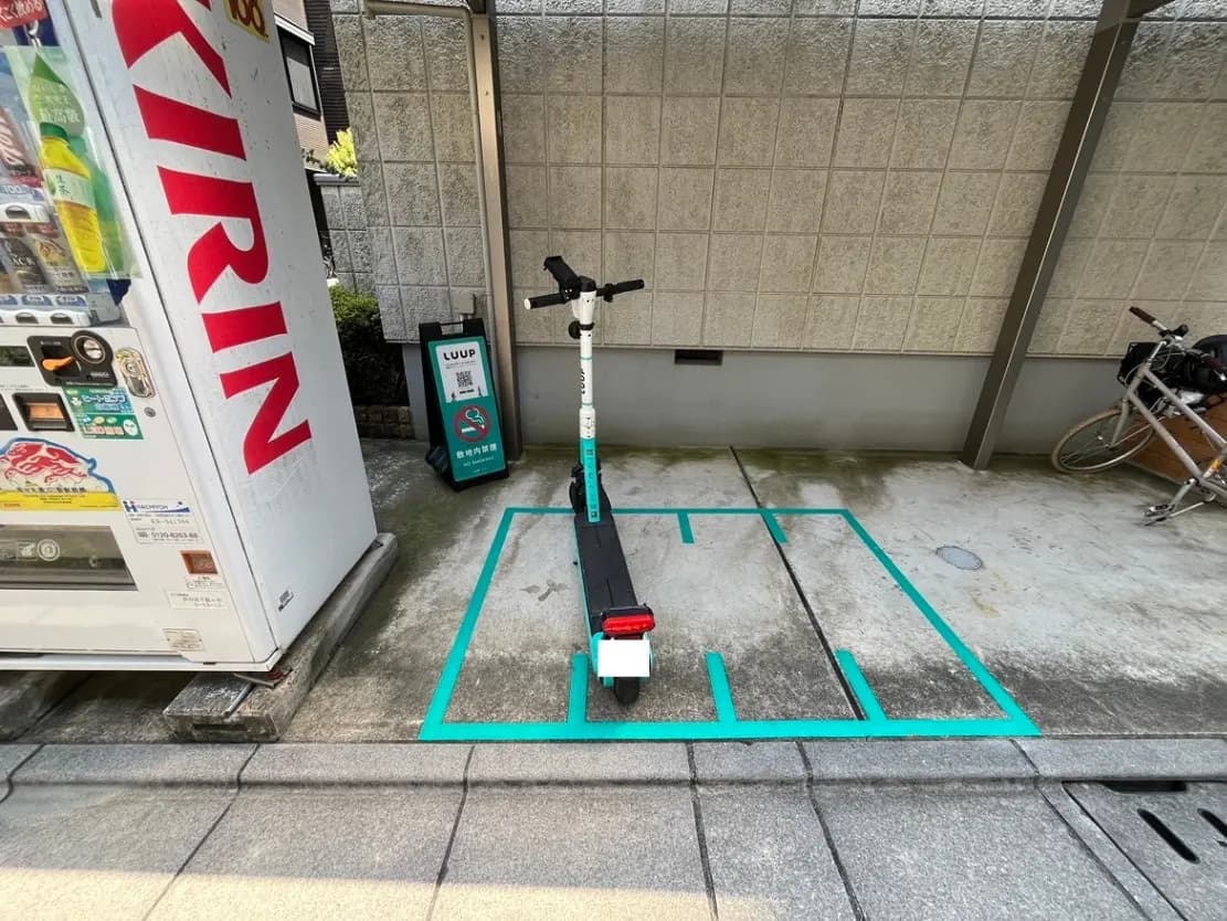 - 自動販売機の横に停められた電動スクーター、青いラインで明示された駐車スペース、周囲には自転車も見える街の風景