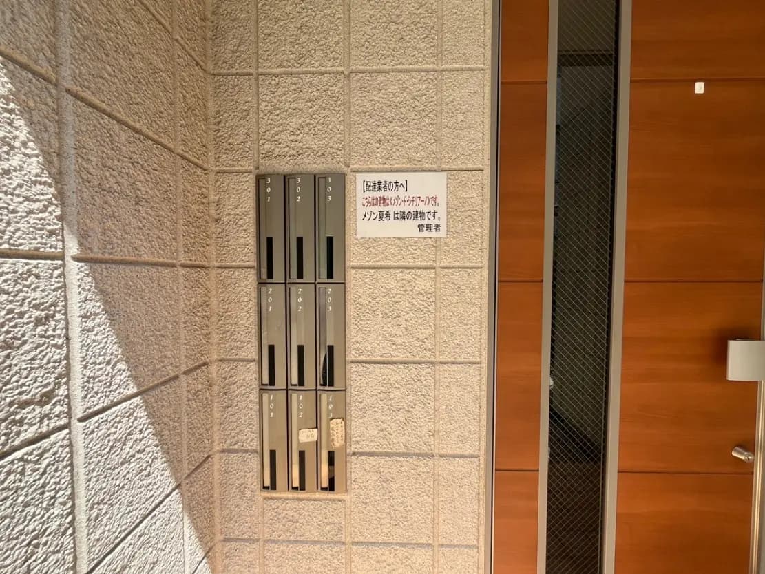 - アパートメントの郵便受け、一覧番号が表示された金属製のボックスと注意書きの貼られた壁の一角