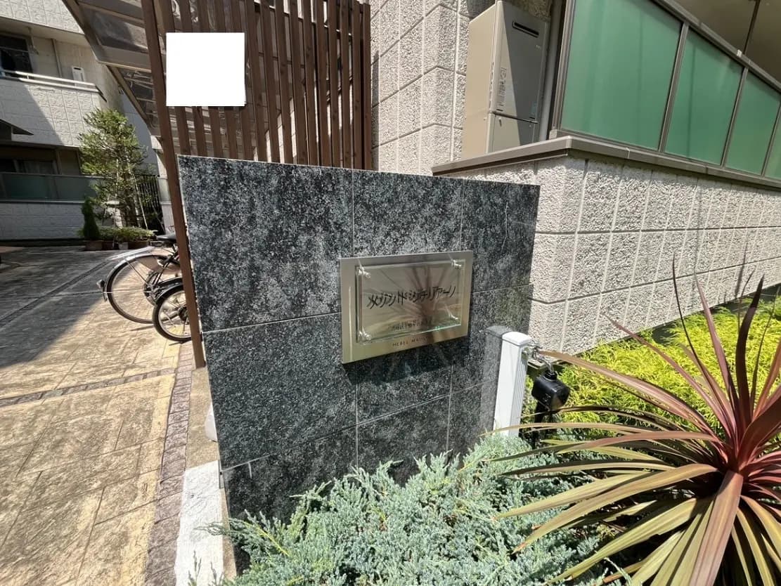- 石材の表札が設置されたマンションのエントランス、周囲には植栽と自転車置き場がある外観