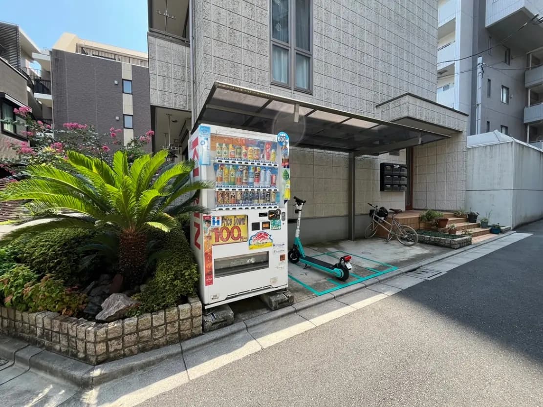 - 建物の横に設置された自動販売機と青い電動スクーター、周囲には緑の植物が植えられた静かな街角の風景