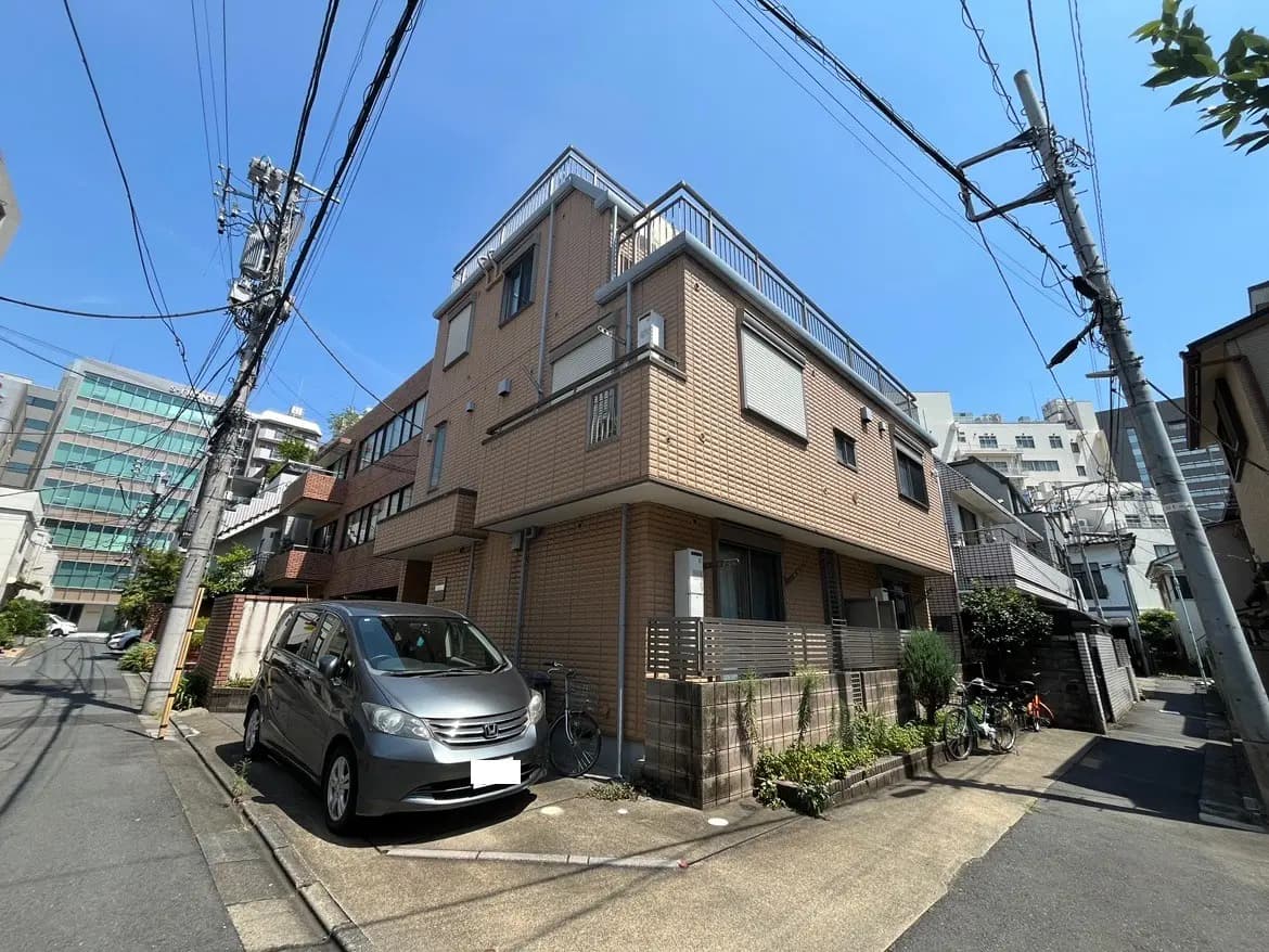 - 都市の住宅街にあるレンガ造りのビル、青空と電柱が印象的で、駐車スペースには小型車が停まっている風景