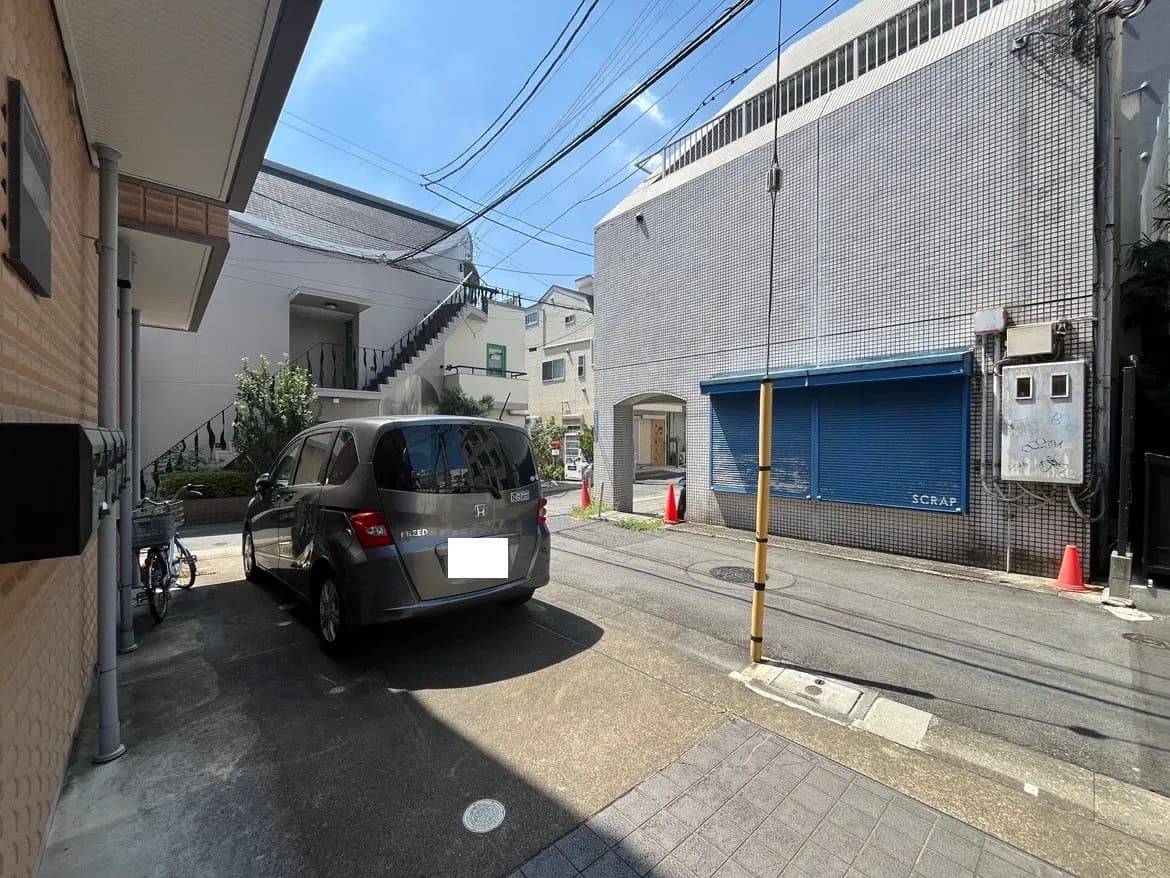 - 駐車場に停められたグレーのミニバン、周囲には住宅や店舗が立ち並ぶ都市の風景、晴れた青空が広がっている景色
