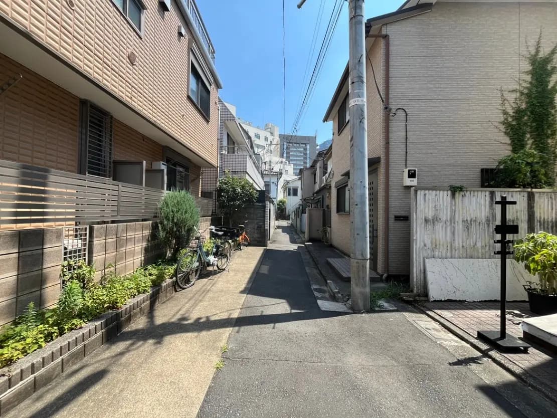 - 住宅街の狭い路地、日差しの中に自転車が停まっており、周囲には緑や壁のデザインが見える風景