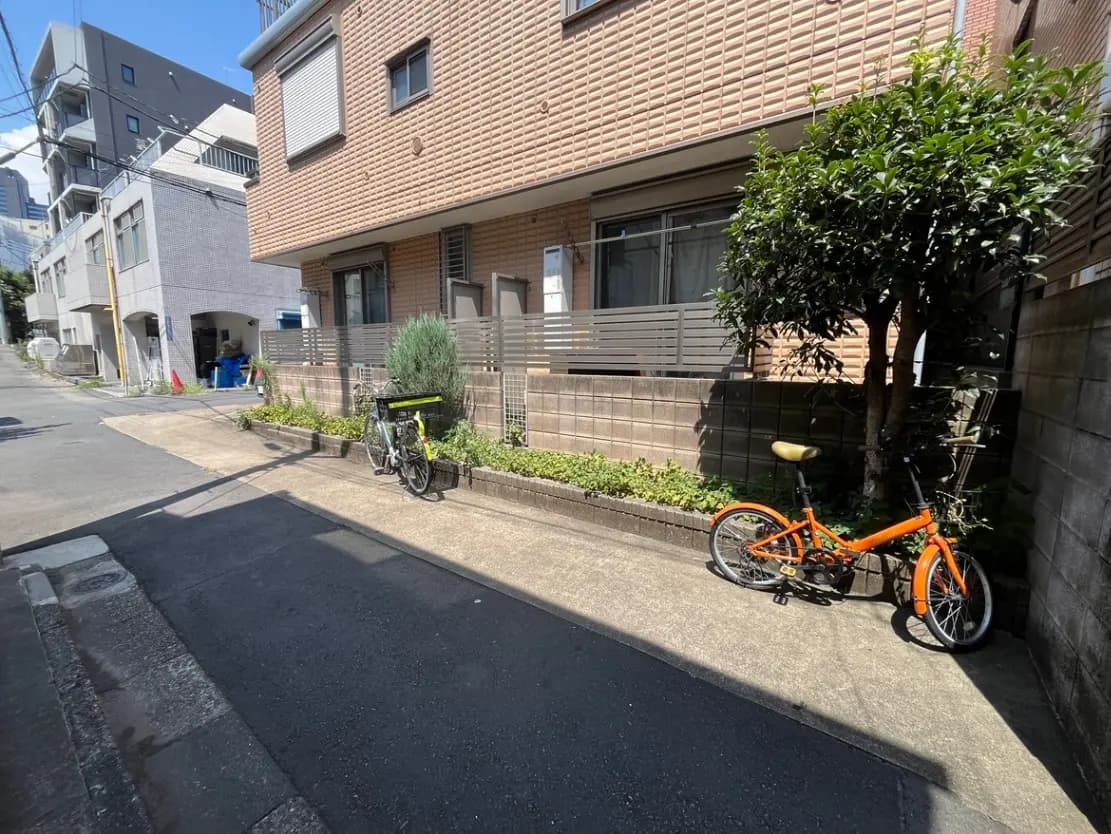 - 住宅街の歩道沿いに停められたオレンジの自転車と緑豊かな植栽の風景、背景に現代的な建物が見えるシーン