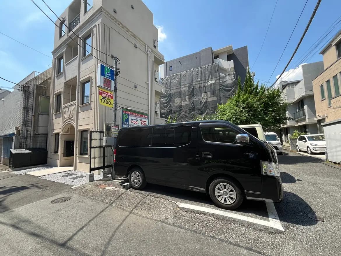 - 駐車場に停まる黒いワンボックスカーと、周囲に立ち並ぶ近代的な建物、晴れた空が広がる街並みの風景