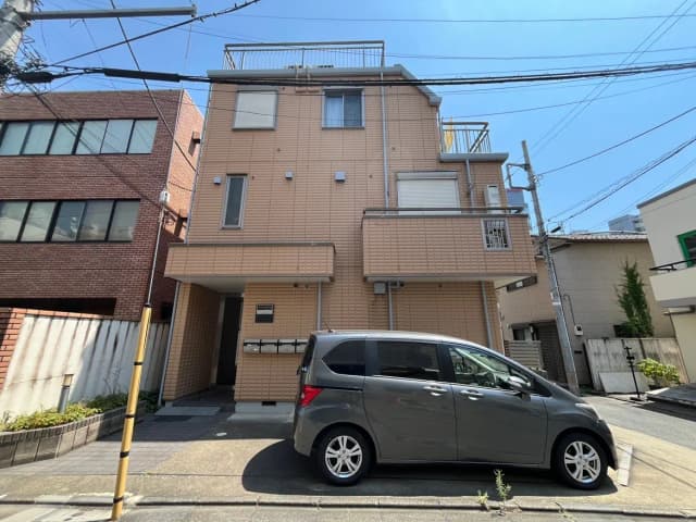 - モダンな外観のアパートメントとその前に駐車されているグレーのコンパクトカー、晴れた青空の下での街の風景