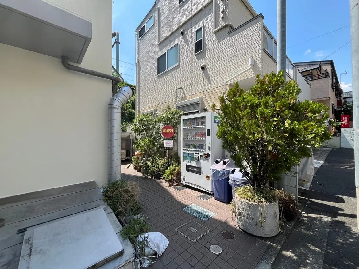 - 硬貨式自動販売機が設置された住宅の脇道、植物やごみ箱がある清潔な街並みの風景