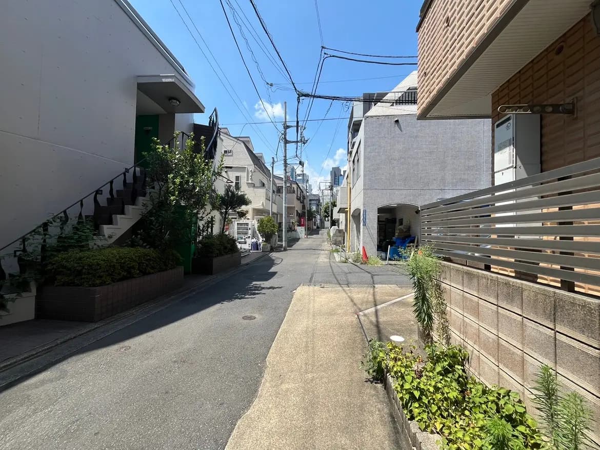 - 青空の下、緑に囲まれた住宅街の細い路地、階段と植栽のある立体的なアプローチが特徴の住宅風景