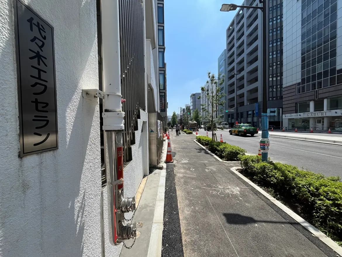 - 代々木エクサロンの看板と周囲の都市風景、整備された歩道に沿って並ぶ高層ビルと緑豊かな植栽の様子