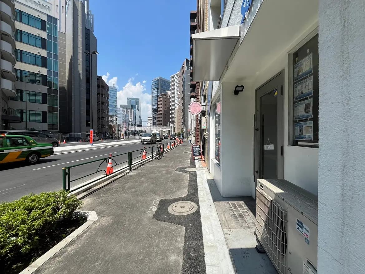 - 都市の街並みと青空、道路に並ぶ車両と施工中の交通標識、店舗の入り口と冷房機が見える風景
