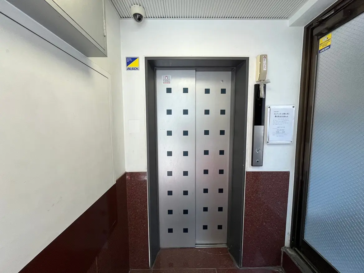 - 銀色のエレベーターの扉があるビル内の風景、隣にはインターホンと案内板が設置されたエントランスエリア