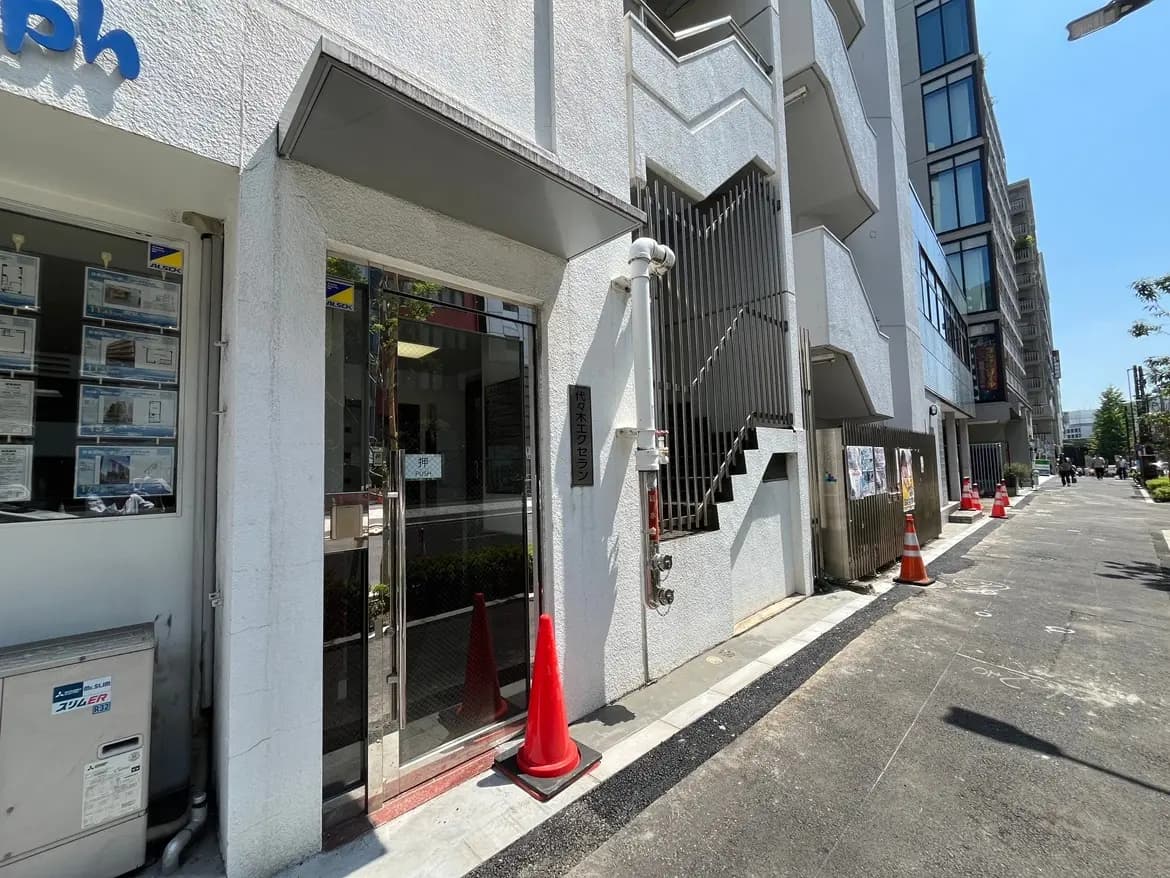 - 現代的な建物の入口、ガラスドアと業務案内の掲示板、周囲に赤いコーンが配置された道の風景