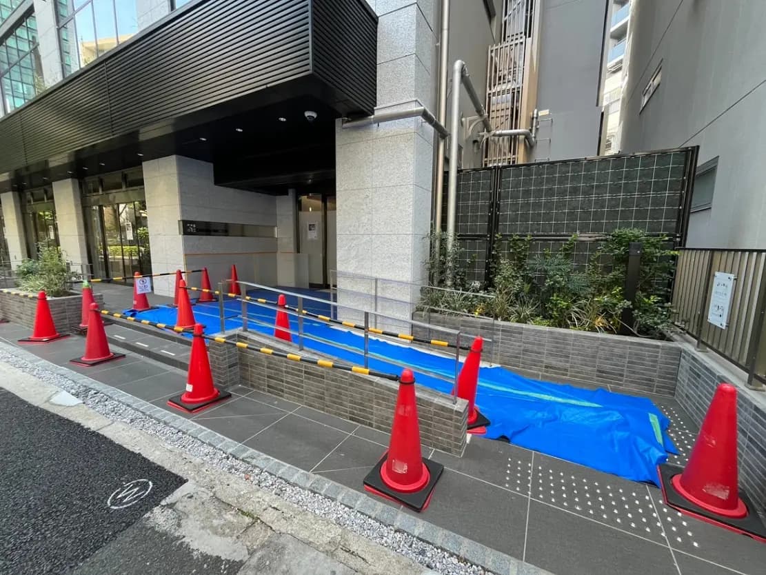 - 建物入り口のバリアフリー用スロープ、交通安全用コーンと青いシートが設置された歩道の様子