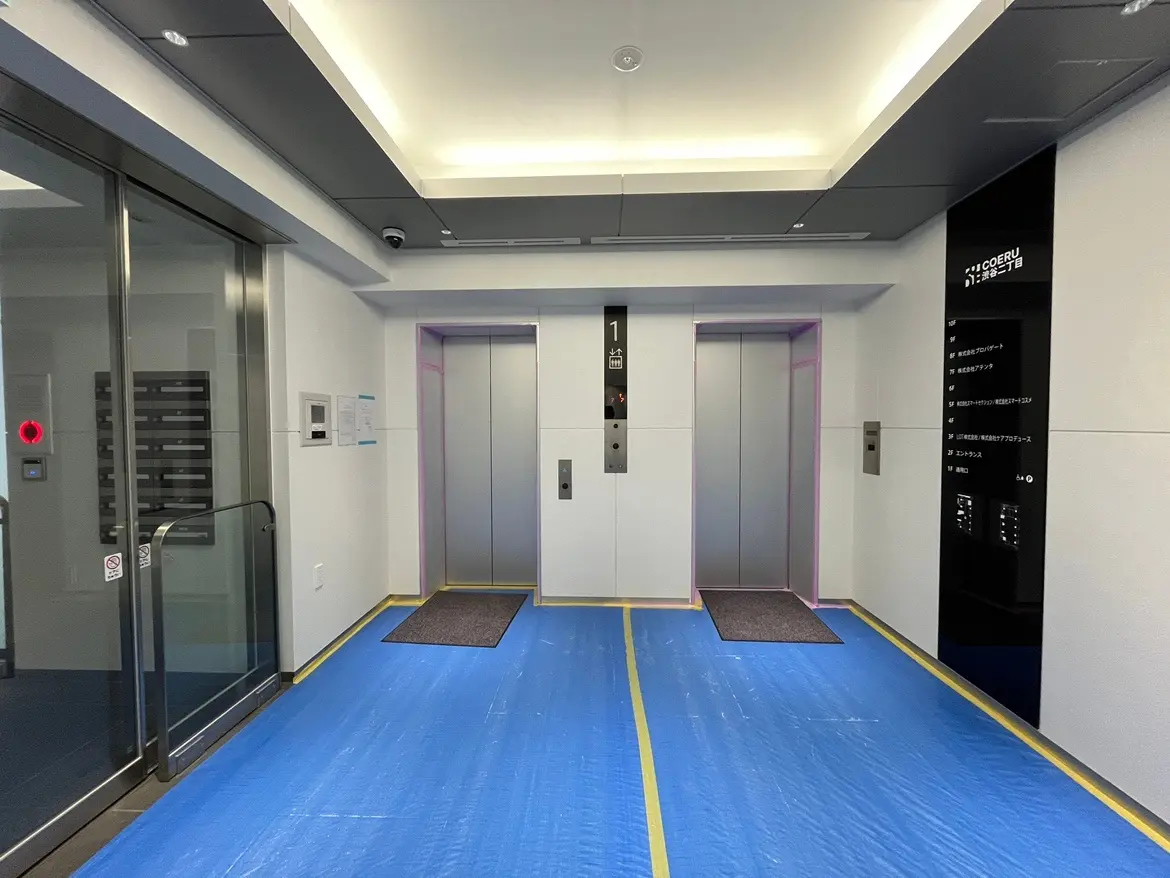 - 複数のエレベーターがある現代的なビルのエントランス、床にはブルーの養生シートが敷かれ、照明が明るいインテリア