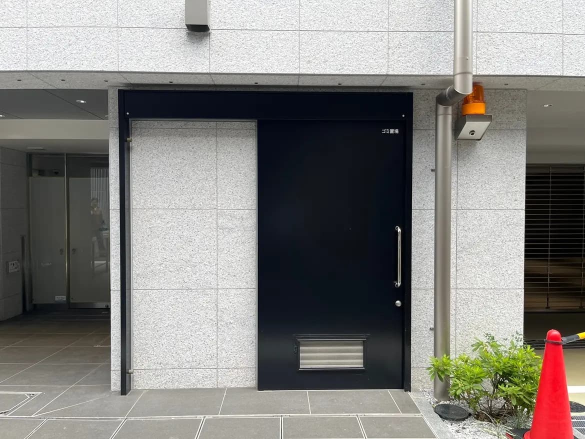 - 黒いドアとグレーのタイル張りの外壁、隣には駐車場の目印となるオレンジのカラーコーンが置かれた建物のエントランス