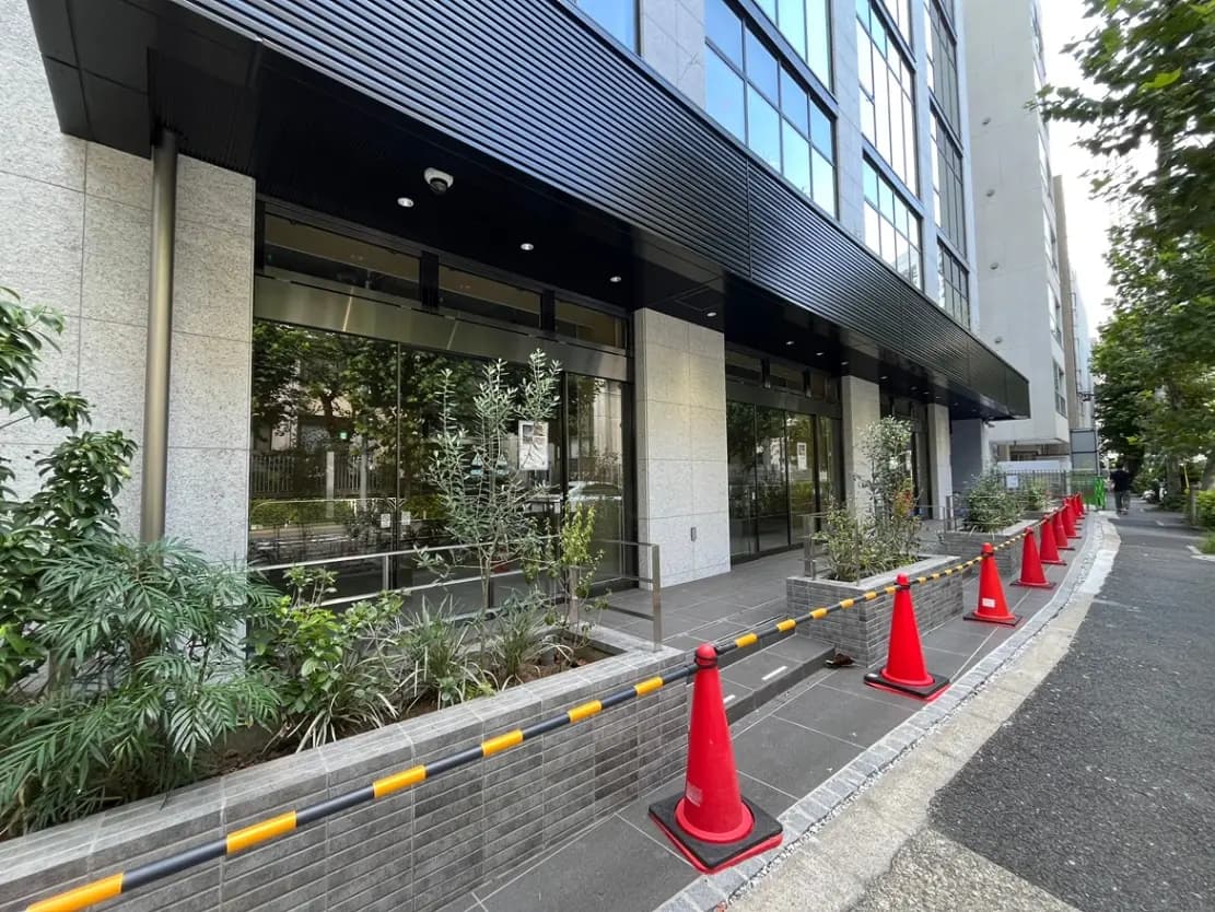 - 現代的なビルのエントランス、周囲に植栽が施され、赤いコーンで囲まれた安全対策の風景