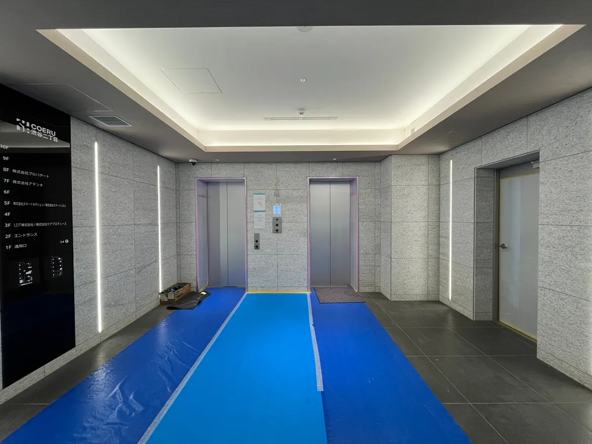 - 現代的なデザインのエレベーターホール、グレーの石材壁、明るい照明とブルーの養生シートが敷かれた床のインテリア