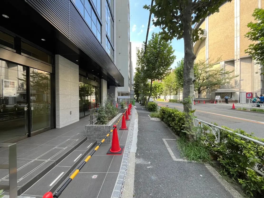 - 近代的なビルのエントランスと歩道、赤いコーンが配置されている街並み、緑の木々と整備された植物の風景