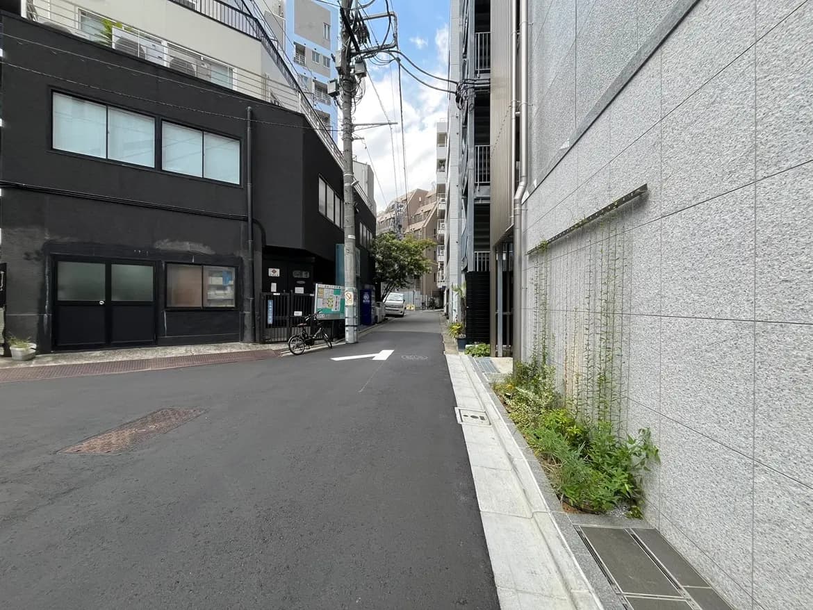 - 都市の狭い通り、左側に黒い外壁の建物、右側にはグレーのタイル張りの建物と緑の植栽が見える風景