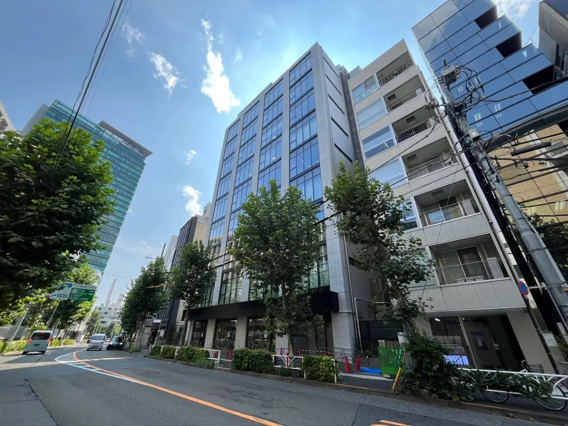 - 高層ビルが立ち並ぶ都市の風景、緑豊かな街路樹と晴れた青空、左側に見える車道と停車中の車両