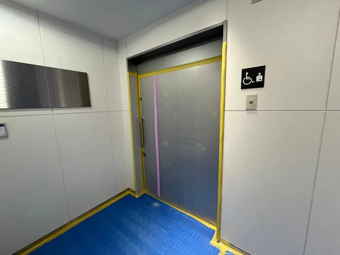 - 車椅子マークのあるバリアフリー対応のエレベーター入口、周囲には養生テープが施された白い壁と青い床の環境