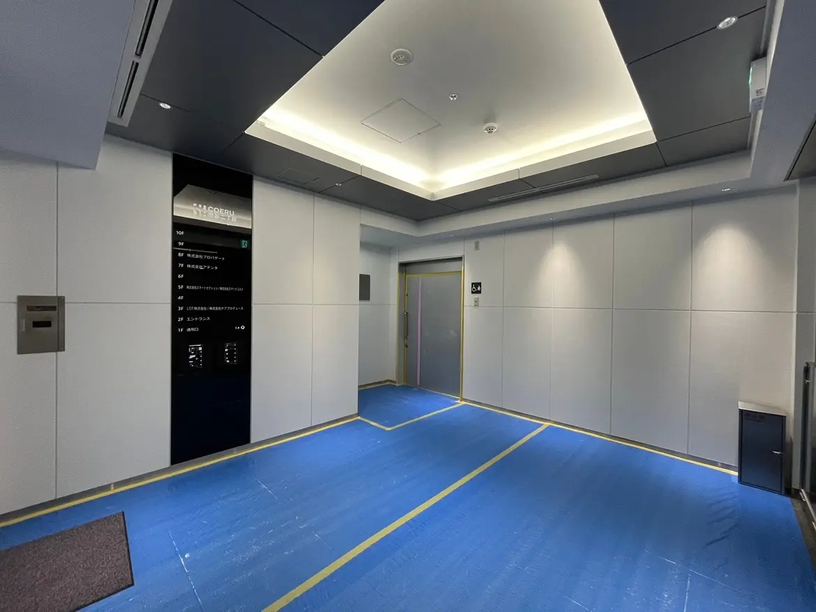 - モダンなデザインのエントランスホール、青い養生シートの床、壁に設置された階数表示板とエレベーターのドアがあるインテリア
