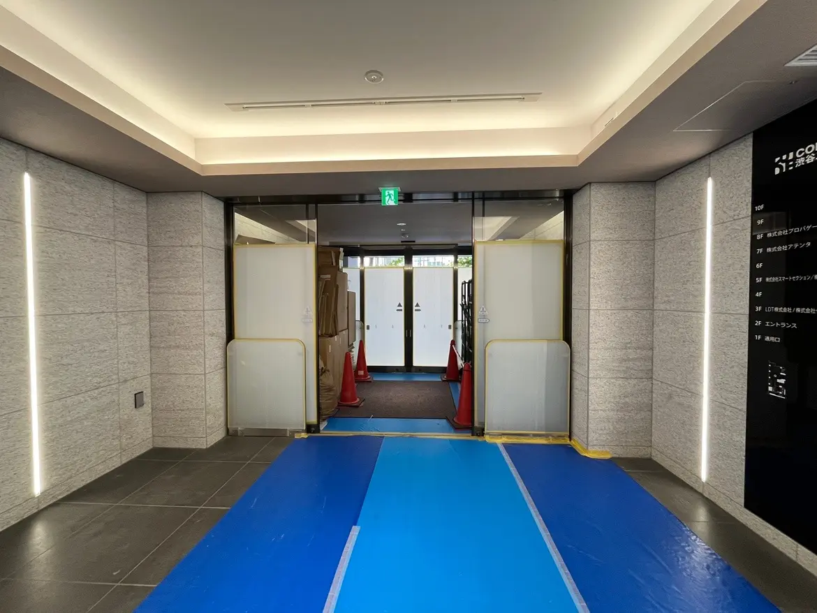 - 商業ビルのエントランス、青い養生シートが敷かれ、周囲には工事用のコーンと段ボールが並ぶ現場の様子