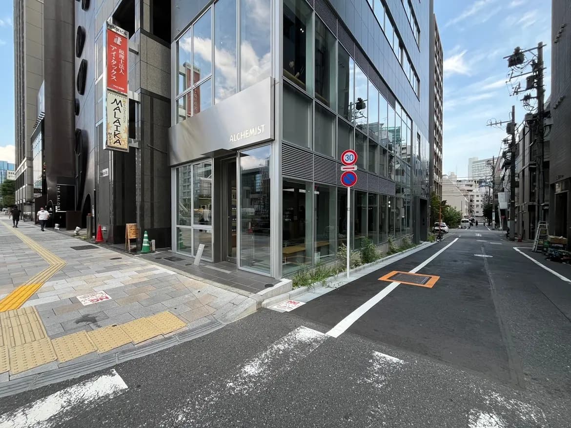 - モダンなカフェ「ALCHEMIST」の外観、横の街並みと歩行者、自転車が通る日常的な風景