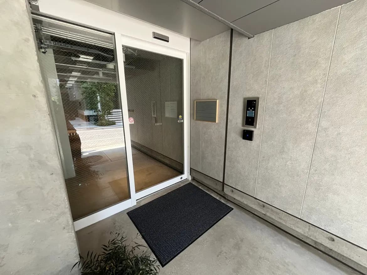 - シンプルでモダンなデザインのエントランス、ガラスの引き戸とタイル張りの床、インターホンと案内プレートが壁に設置された様子