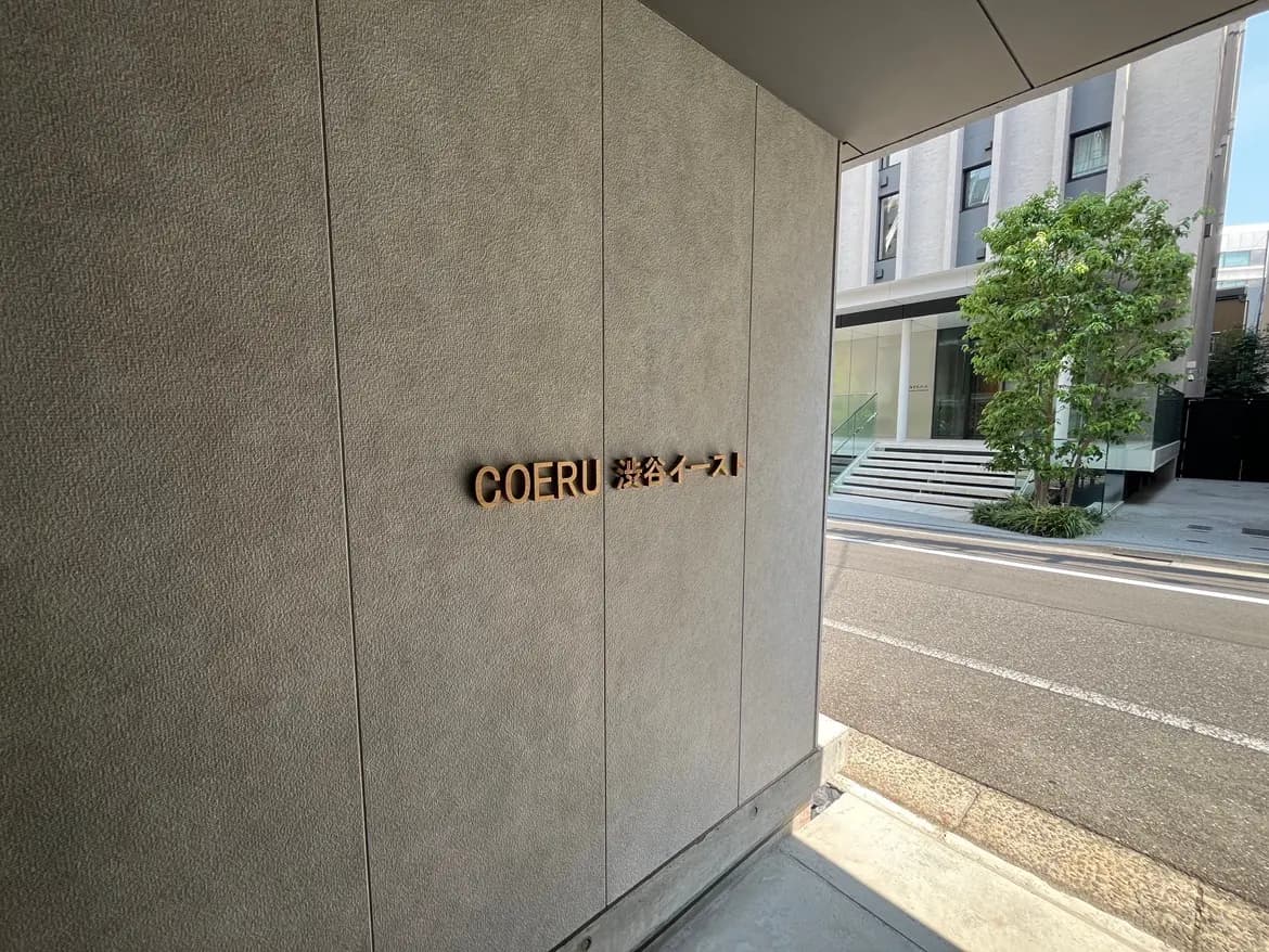 - 「COERU」と書かれた木製のサインボードが取り付けられた現代的なエントランス、周囲には緑の木々が見える景観